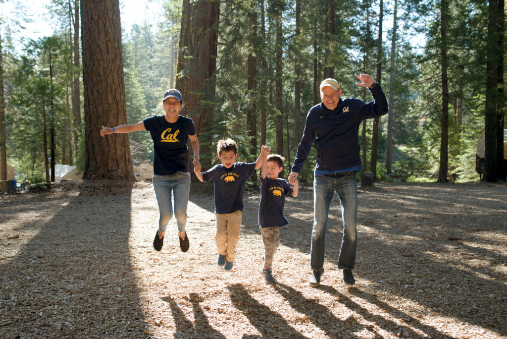 Family wearing Cal shirts photographed in mid-air while holding hands and jumping in the woods