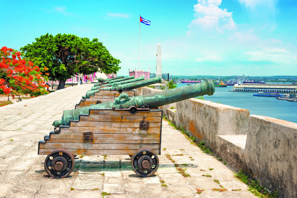 Rows of historic cannons on wooden carriages at La Cabana Fortress overlook a vibrant harbor with a Cuban flag fluttering in the background.