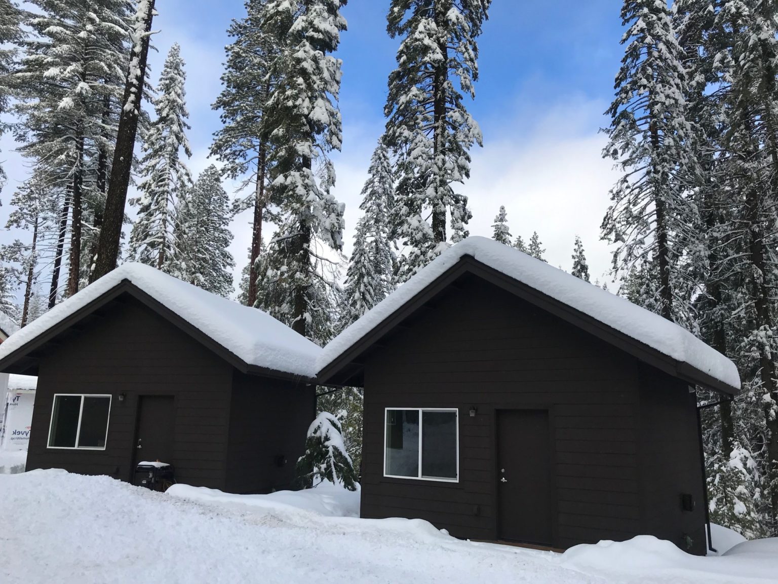 2 new winterized cabins at Camp Oski, which are dark brown, covered in snow, and nestled in a backdrop of snowy pine trees and blue skies.
