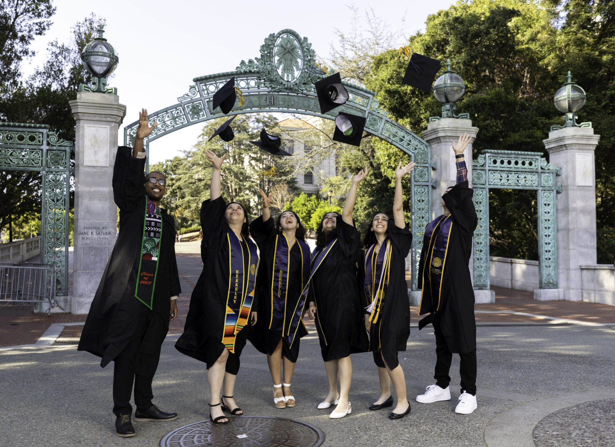 A group of scholars dressed in graduation gowns throw their graduation caps into the air in front of Sather Gate.