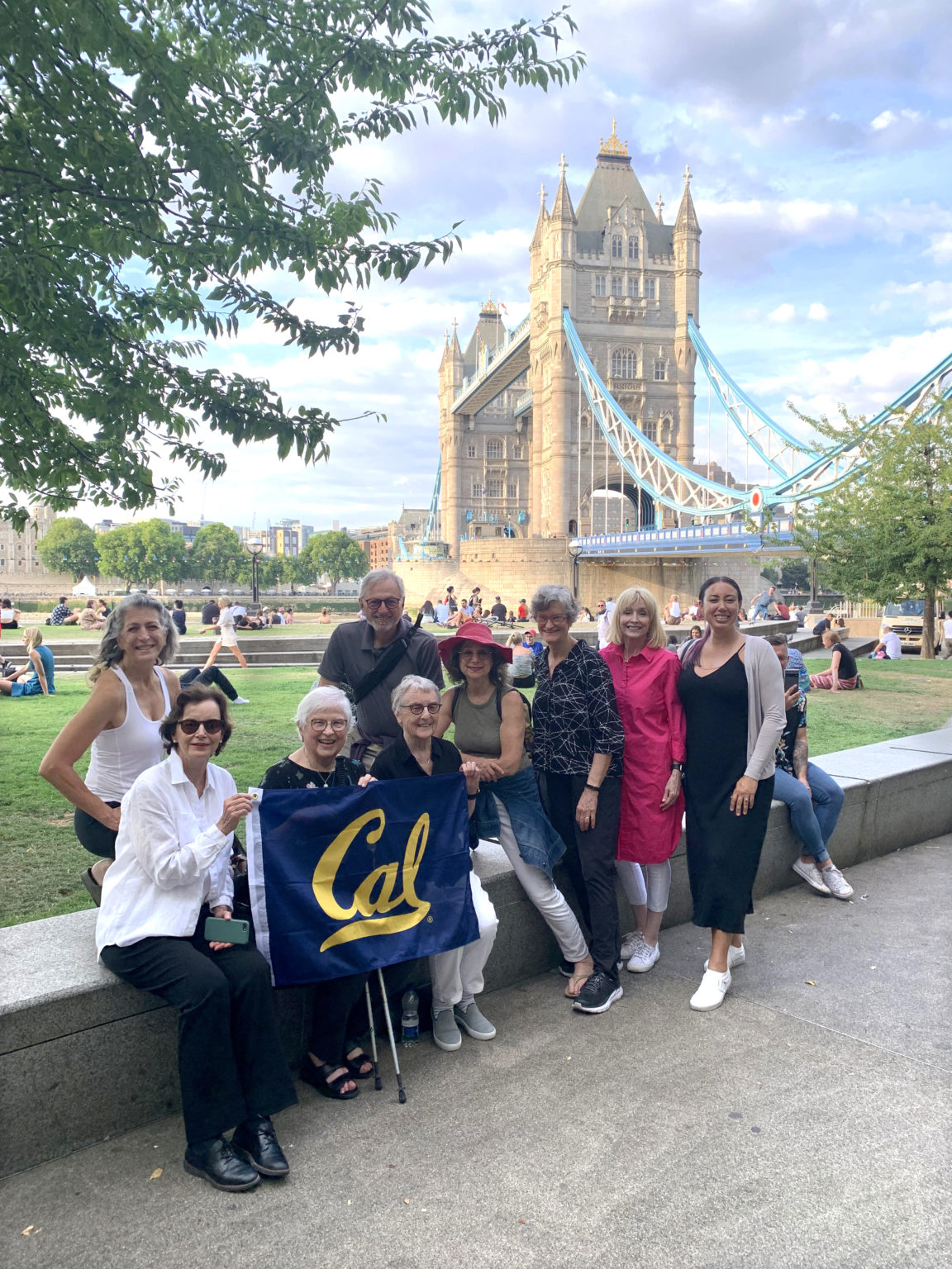 A group of 9 people holding a Cal flag and posing in front of the London bridge.