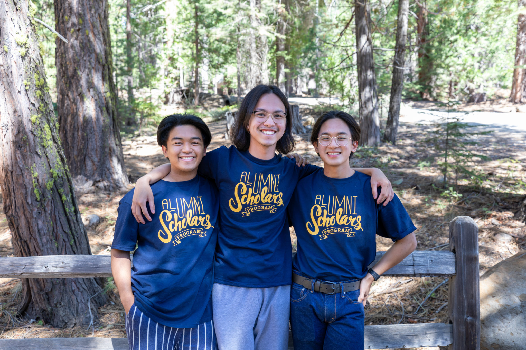 Three students smiling with their arms around each other with woods in the background. They are all wearing Alumni Scholars Program T-shirts.
