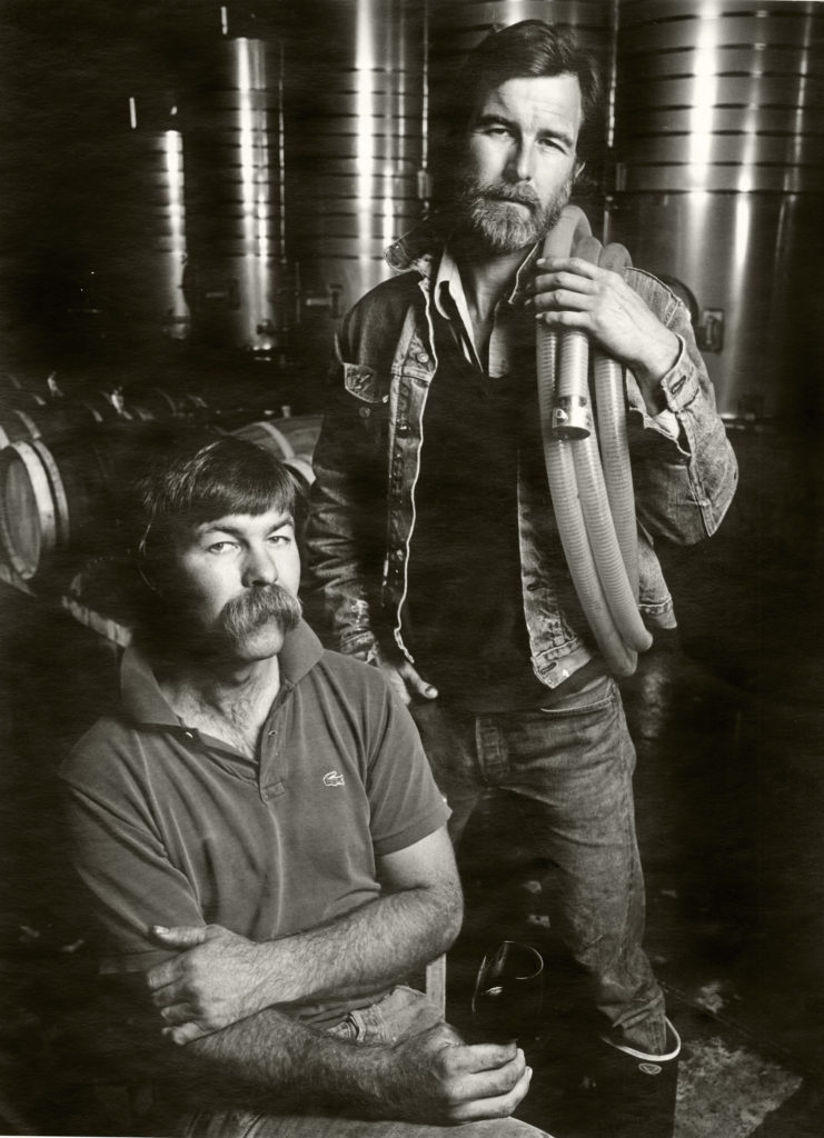 stuart smith stands behind his brother charles in front of wood and steel wine barrels