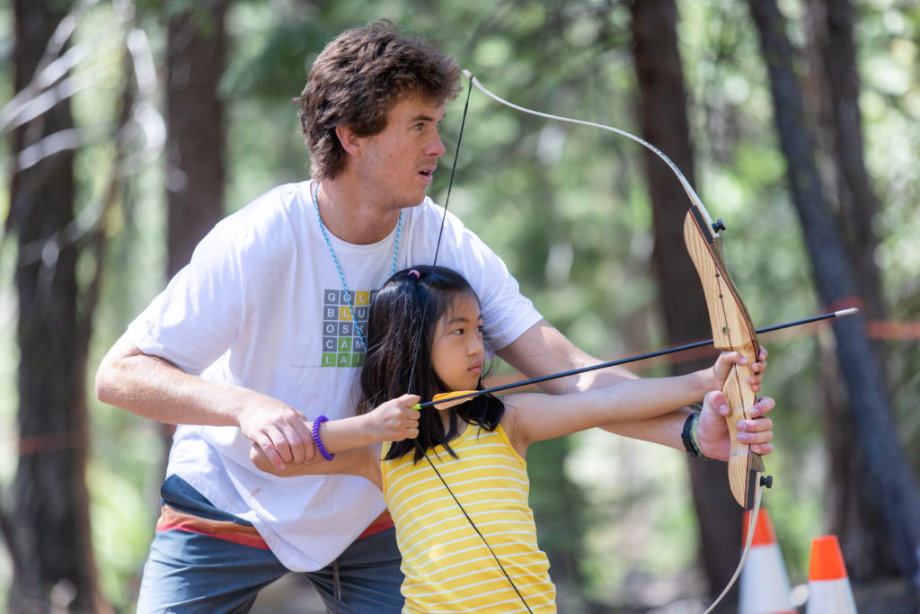 A Lair staffer instructing archery to a younger girl with her bow raised.