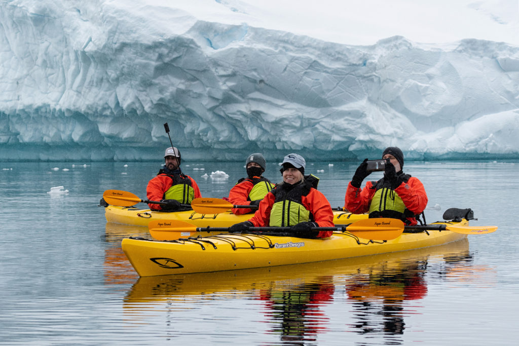 Four guests in orange wetsuits in yellow kayaks on open water with iceberg behind them