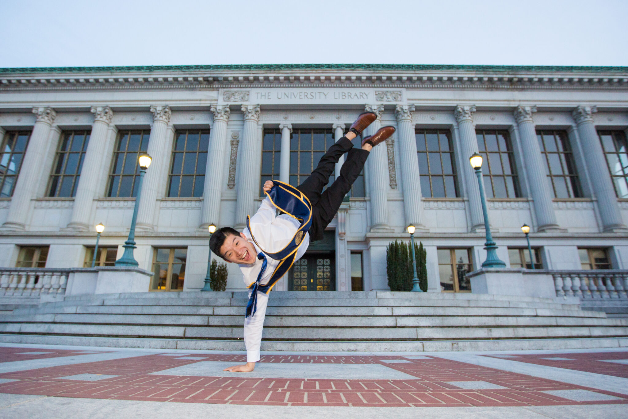 Robert Yu performs a dance move in front of the Bancroft Library wearing his graduation stole.