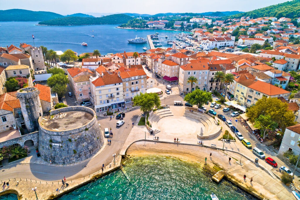 Aerial view of a coastal town with terracotta roofs, palm trees, a harbor, and a stone tower, surrounded by lush hills and turquoise water.