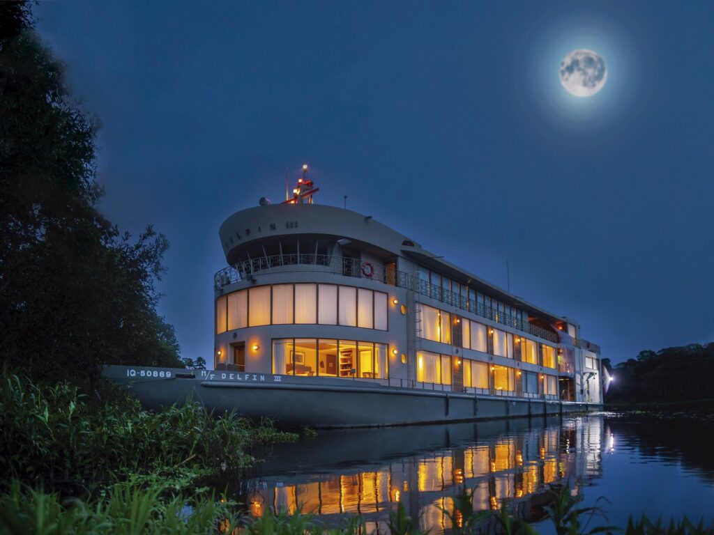 The Delfin III ship floats on the Amazon River at night under a luminous full moon