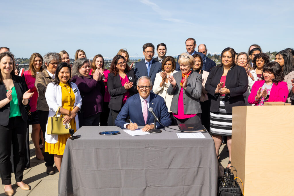Jay Inslee smiles while signing a bill with lots of people around him clapping