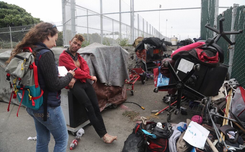 Raguso interviews someone in what looks like a homeless encampment