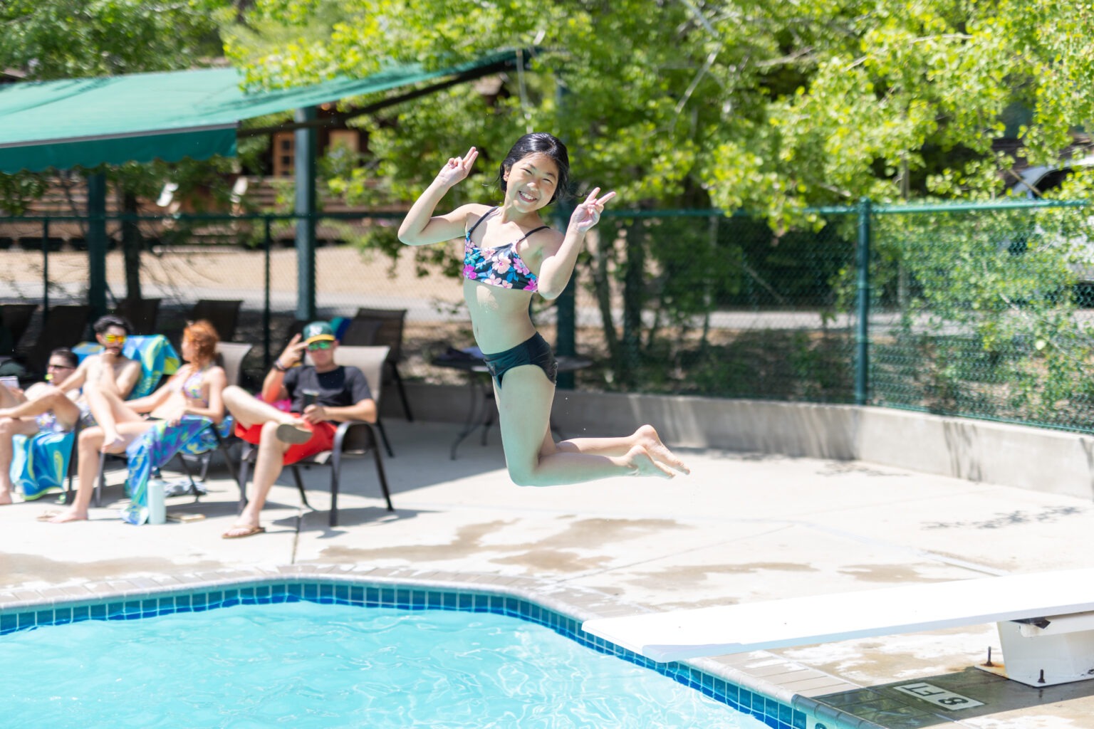 young girl jumping off the diving board smiling at the camera and putting up peace sign fingers