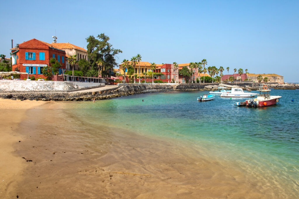 Coast of Goree Island with small boats in water and red and yellow buildings along the shorline