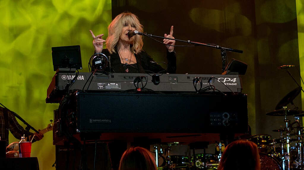 Christine McVie, the keyboardist and vocalist of Fleetwood Mac, is performing on stage. She is playing a Yamaha CP4 stage piano and singing into a microphone.