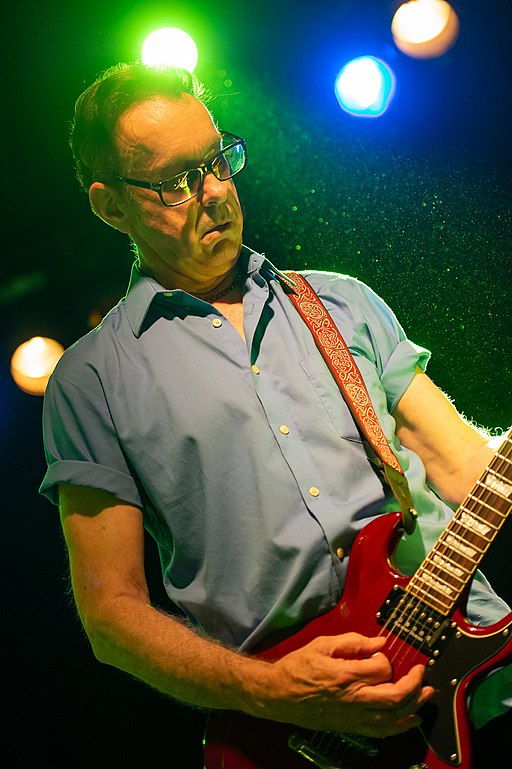 East Bay Ray is performing on stage. He is playing a red electric guitar and is dressed in a light blue shirt with rolled-up sleeves.