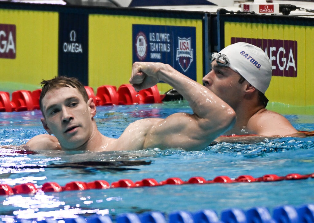 Swimmer Ryan Murphy celebrates in the pool, flexing his arm after a race, with another swimmer in the background wearing a white cap labeled "Gators." 