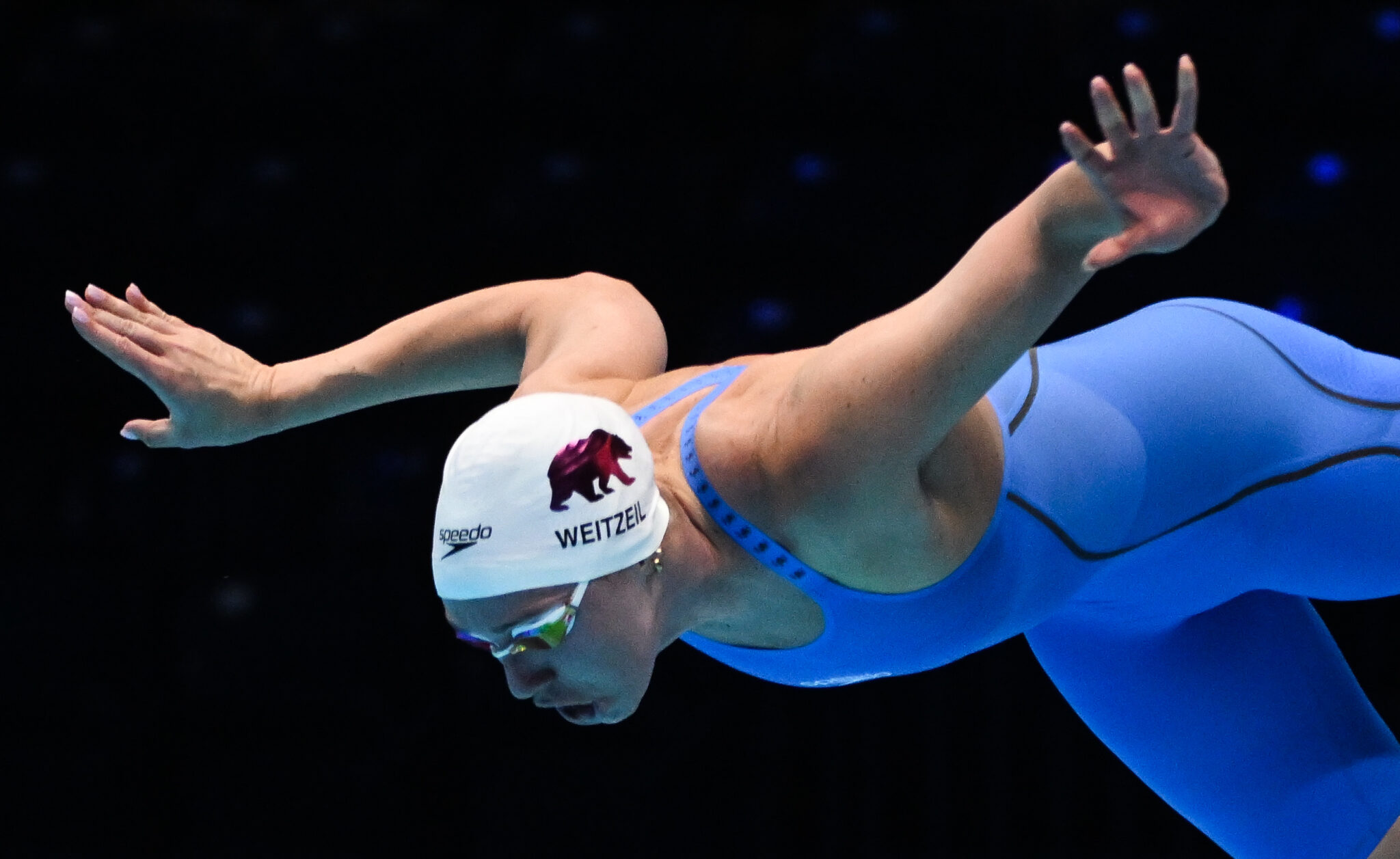 Swimmer Abbey Weitzeil, wearing a white swim cap and blue swimsuit, dives into the water with arms outstretched.