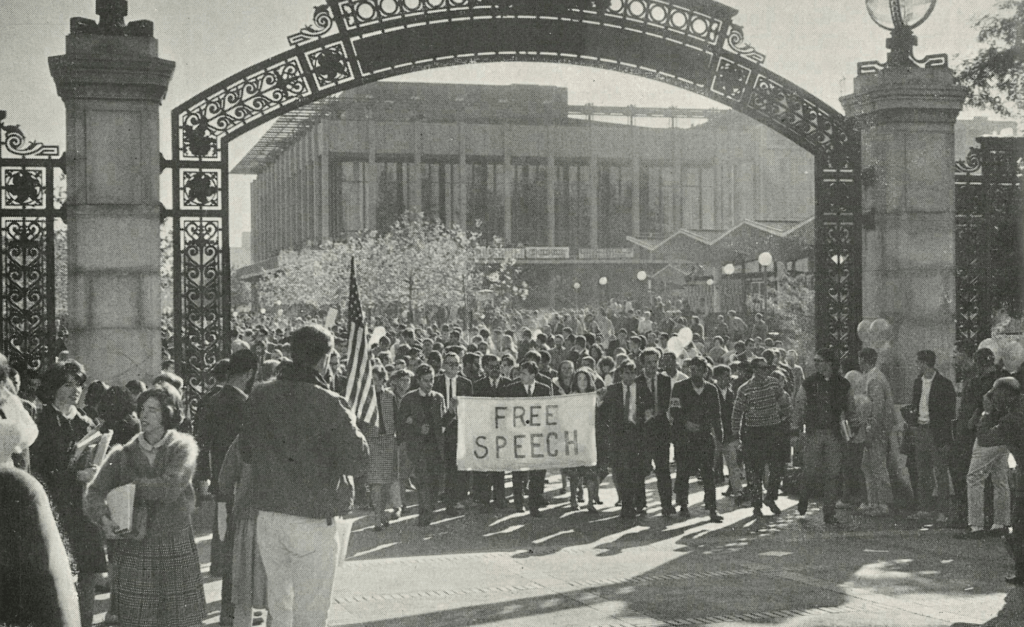 Black-and-white photo of protesters marching through Sather Gate/ The crowd is carrying a banner that reads 