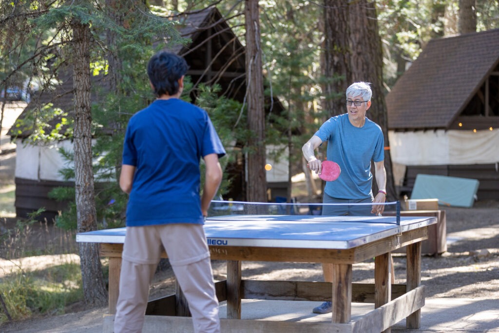 Two men playing ping pong with tents and trees in the background.