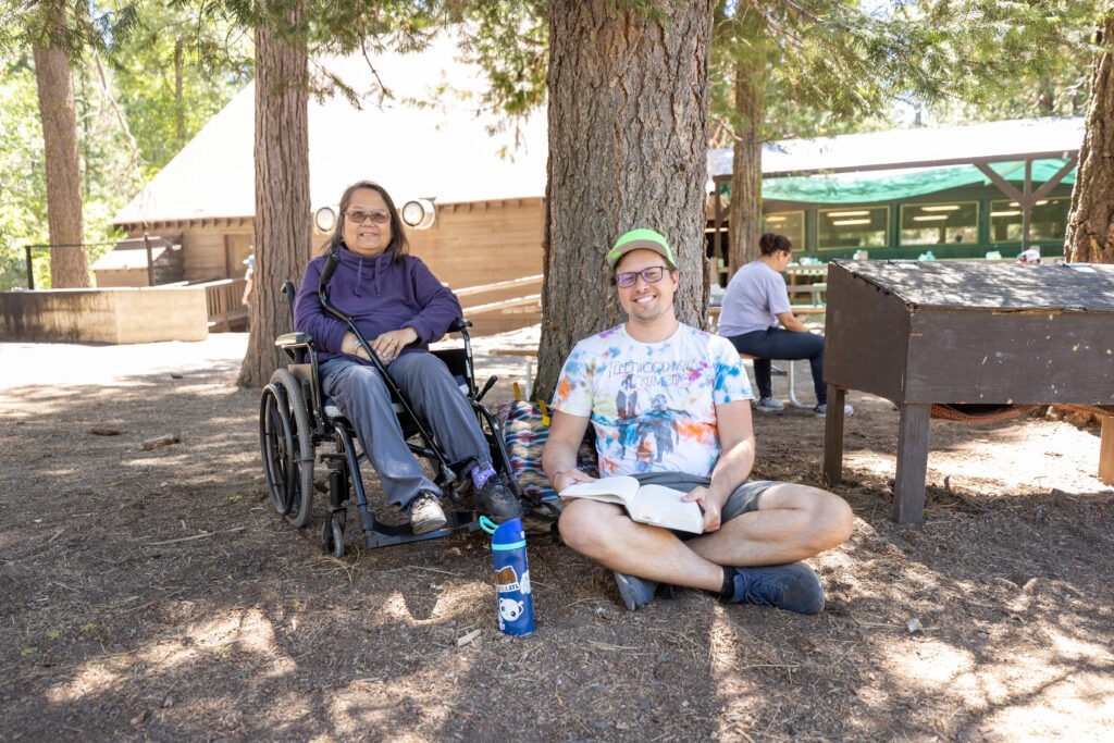 A man sitting on the ground with a book, and a woman in a wheelchair, sitting under trees and smiling.