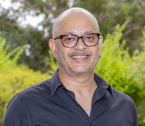 headshot of man in front of bushes wearing glasses, earrings, and black collared shirt