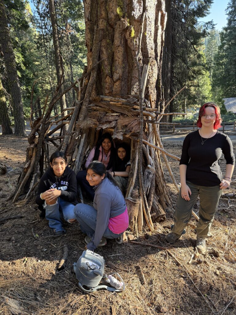 5th grade Oski Science Camp students sitting in and standing next to an outdoor shelter they made using branches.