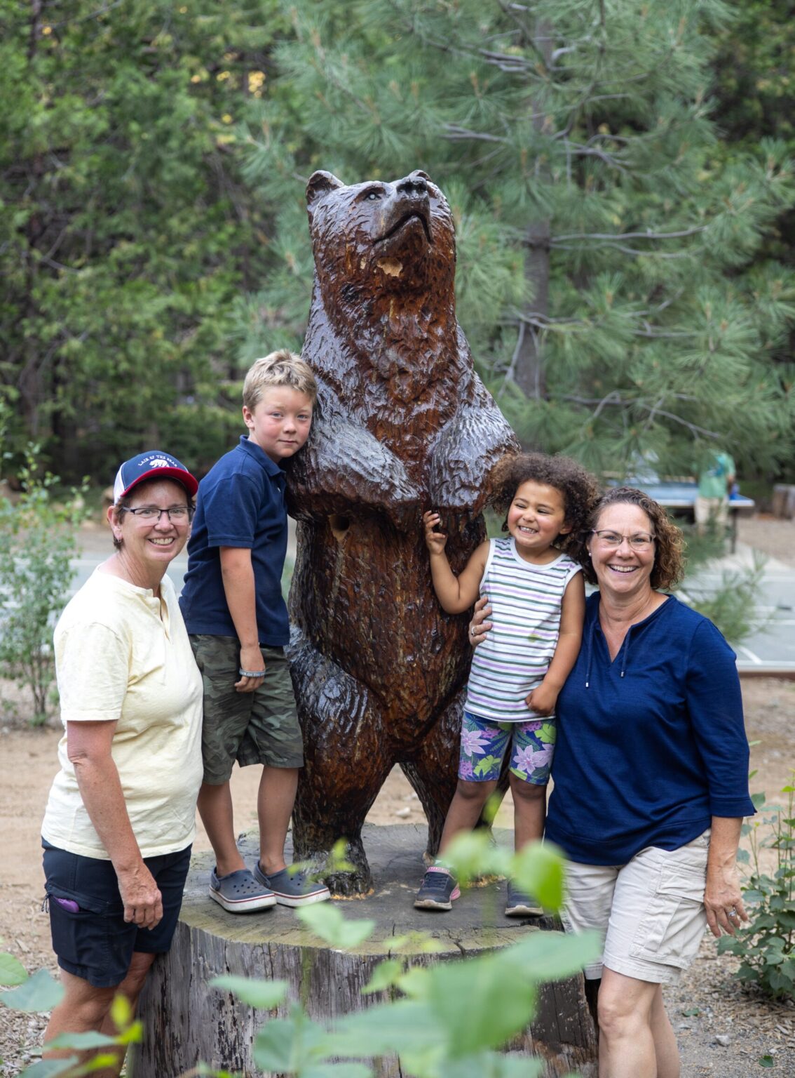 Two moms and two young kids standing smiling with the tall wood carved bear at the Lair
