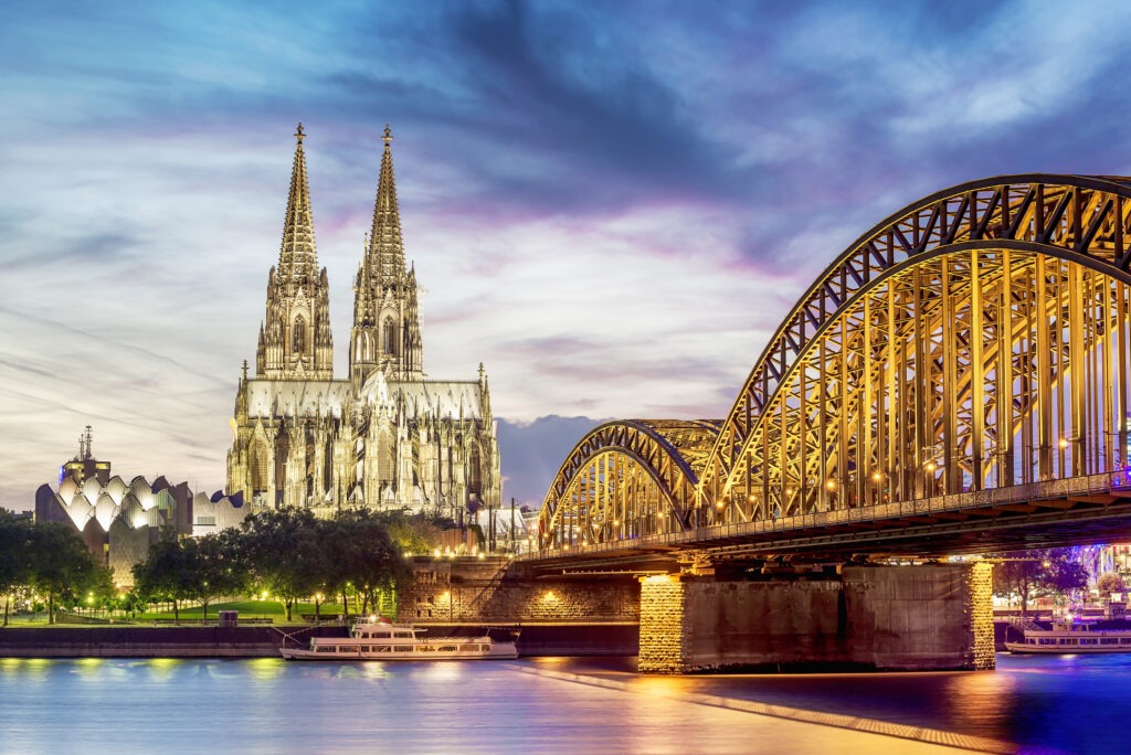 Illuminated Cologne Cathedral towers behind the Hohenzollern Bridge, reflecting in the calm river at dusk