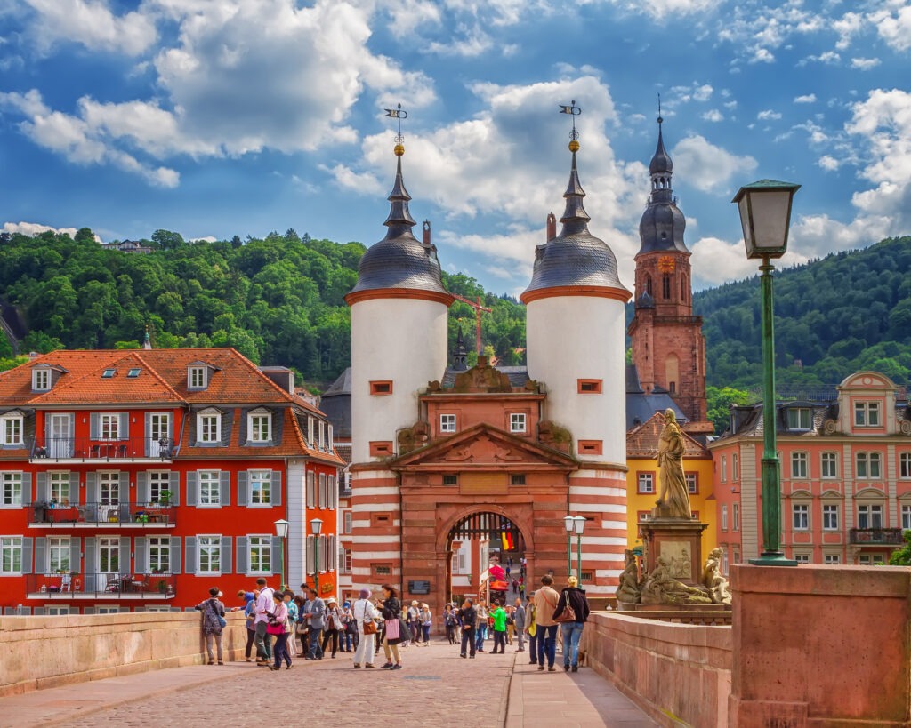 Historic Heidelberg Bridge entrance with striped towers, colorful buildings, and tourists under a scenic sky in a lush green landscape