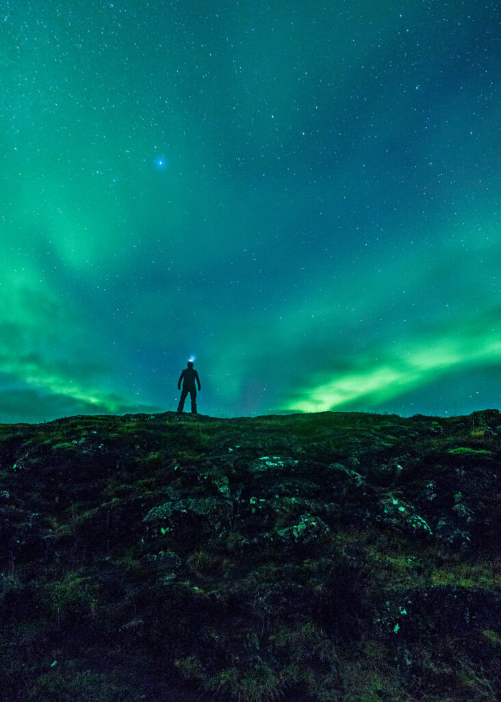 A person standing on a hill with the glowing green northern lights in the sky
