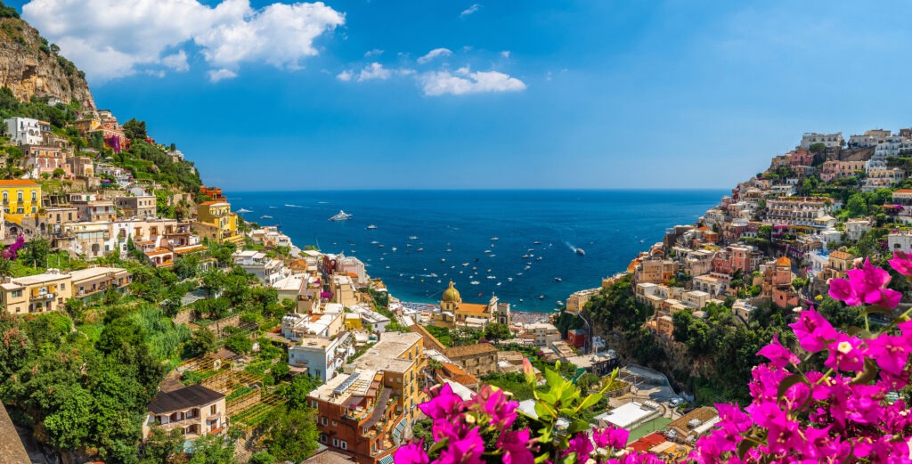 A vibrant coastal view of Positano, Italy, showcasing colorful cliffside houses, lush greenery, and a serene blue sea dotted with boats