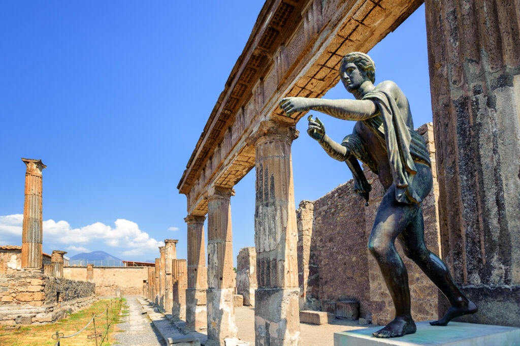 A bronze statue of a figure gesturing amid ancient ruins, with stone columns beneath a clear blue sky in Pompeii.