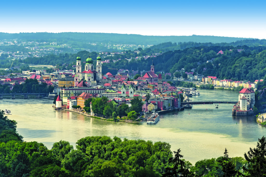 A scenic view of the town of Passau, Germany, with colorful buildings, a river, and churches amid lush green hills.