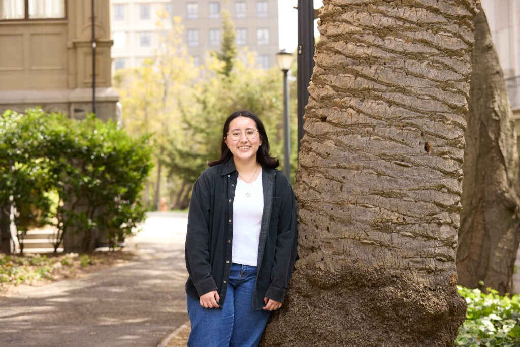 Melissa Mora-Gonzalez standing beside a textured tree trunk on the campus.